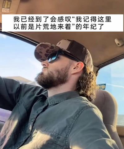 这是一张带有文字注释的静态真人图片，图中一位戴着迷彩棒球帽、墨镜，留着络腮胡和鲻鱼头的男子坐在汽车驾驶座上，神态略显沧桑。图片上方配有文字“我已经到了会感叹‘我记得这里以前是片荒地来着’的年纪了”，表达了人在年龄增长后，见证周边环境变迁，生出岁月流逝、物是人非的感慨，这种感慨能引发不少人的共鸣，是对自身成长和时光变化的自嘲式感叹。