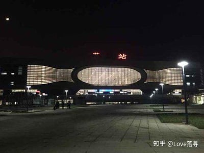 这是一张夜晚拍摄的现代化火车站照片，车站建筑外观设计极具现代感，正面拥有大面积的玻璃采光结构与灯带装饰，顶部红色的“站”字标识醒目，左侧的完整站名文字被遮挡。站前广场空旷开阔，数盏路灯提供照明，广场上有零星行人活动，整体氛围静谧，展现了夜间火车站的冷清与规整的建筑风貌。