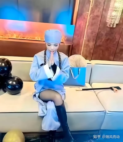 图中是一位穿着浅灰色尼姑服饰的女性，戴着同色僧帽、挂着佛珠，双手合十做出祈祷的姿势，但下半身搭配了黑丝和黑色长靴，形成强烈的风格反差。她坐在现代风格的拼接沙发上，沙发旁放置着类似僧人的道具小锄头和蒲扇，背景是带有蓝橙灯光的室内环境，整体氛围搞怪又充满违和感，通过服饰混搭制造出趣味效果。