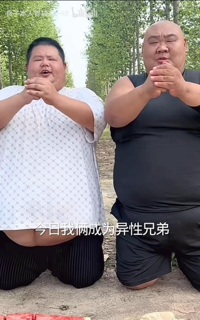 画面中两位体型肥胖的男子在户外树林旁的泥土地上跪地，双手抱拳作揖，模仿传统的结拜仪式，表情认真却带着滑稽感。画面下方的字幕显示“今日我俩成为异性兄弟”，这里是玩了谐音梗，将原本的“异姓兄弟”误写/误说成“异性兄弟”，制造出荒诞搞笑的效果，整体场景充满了接地气的网络整活氛围。