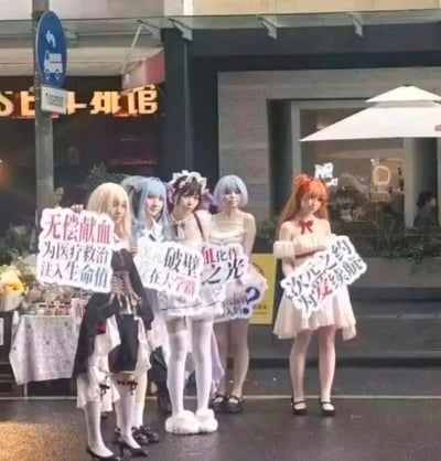 图片展示了五位身着二次元角色cosplay服装的女生站在街头，开展无偿献血的公益宣传活动。她们手持的宣传牌文案巧妙结合了二次元文化梗与公益主题：“无偿献血 为医疗救治注入生命值”借用游戏中“生命值”的概念，类比献血能为患者延续生命的意义；“破壁 在大学路”中的“破壁”指打破次元壁，寓意让二次元文化与三次元公益活动融合，地点指向年轻人聚集的大学路；“血化作光？”玩了谐音与双关梗，将“血液”的“血”与“化作光”结合，既点出献血的核心，又传递出公益带来的温暖希望；“次元之约 为爱续航”用“次元”呼应二次元主题，把献血比作“为爱续航”，拉近与年轻群体的距离。背景是街边商铺，整体氛围充满活力，以年轻人喜爱的形式推广无偿献血，降低公益宣传的距离感。