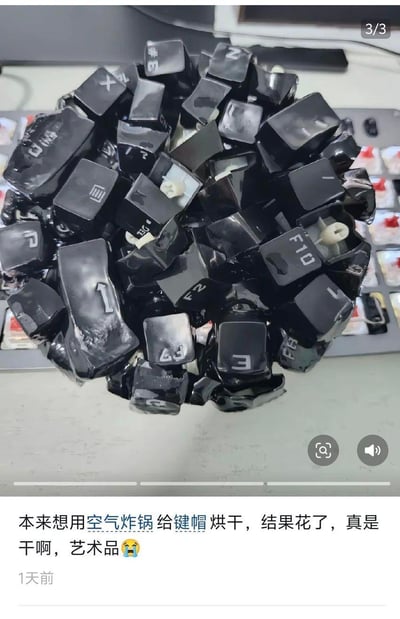 图中展示了因错误使用空气炸锅烘干而被烤变形、融化粘连成球状的键盘键帽，键帽表面的字符因高温变得模糊花掉，背景可见被拆除键帽的键盘。下方是社交平台的用户配文：‘本来想用空气炸锅给键帽烘干，结果花了，真是干啊，艺术品😭’，用户以无奈又自嘲的语气分享了这次操作失误的趣事。