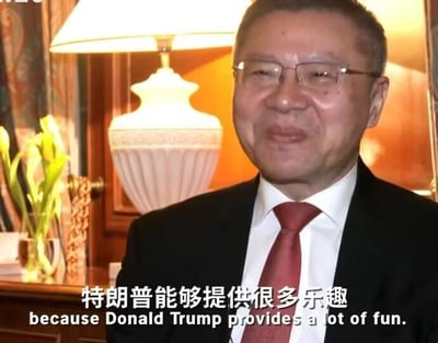 图片中是一位穿着黑色西装、佩戴眼镜的中年男性，面带微笑，背景为室内场景，有一盏白色台灯和装饰花瓶。图片底部有中英文字幕：“特朗普能够提供很多乐趣 because Donald Trump provides a lot of fun.” 整体场景看起来像是在接受采访或进行对话。