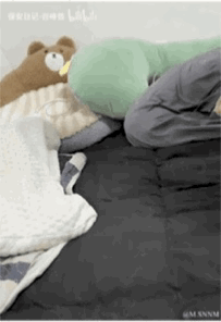 这是一段竖屏动态GIF，呈现了温馨的宅家场景：深色床铺上，有人用被褥将自己遮挡起来，床边摆放着一只呆萌的棕色卡通毛绒熊玩偶和一个绿色U型枕，整体氛围放松又治愈，传递出宅家时的慵懒与惬意。