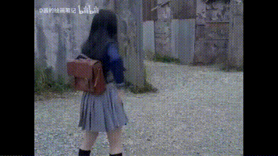 这是一段动态GIF，画面中一位穿着蓝灰拼接校服、背着棕色双肩包的女生背对着镜头，站在铺有碎石的户外区域，身旁有树木和老旧围墙，女生做出即将转身的动作，整体场景带有复古特摄剧的怀旧氛围。