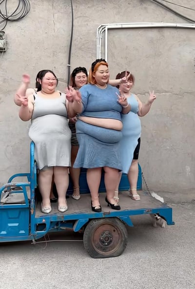 画面中四位体型偏胖的女生站在蓝色电动三轮车的车斗里，她们穿着清凉的裙装，对着镜头开心地挥手、比出V字手势，脸上洋溢着笑容，氛围轻松欢快。右下角有一只小狗从车斗下方探出头来，背景是带有管道和电线的水泥墙面，场景看起来是在户外的居民区小巷中，传递出自信、乐观的生活态度。