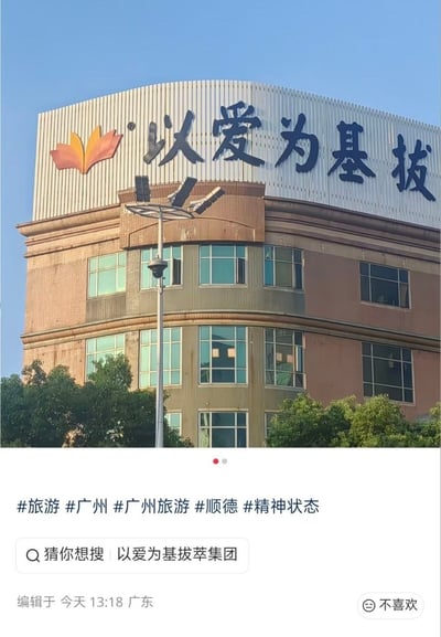 这是一张社交媒体平台的内容截图，主体为广州顺德某建筑的实拍画面，建筑顶部的企业招牌原本完整内容为“以爱为基拔萃集团”，拍摄角度恰好只露出“以爱为基拔”五个字，形成意外的谐音歧义，具有搞笑效果。截图下方可见发布者添加的旅游相关标签，平台自动给出的搜索提示“以爱为基拔萃集团”，以及发布位置为广东、编辑时间为当日13:18的相关信息。