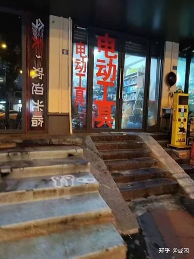 这是一张夜晚拍摄的店铺门口场景图，画面里有两组通往店铺的台阶，左侧台阶的表面上喷绘了两个类似电动工具扳手的图案；店铺的玻璃推拉门上贴着醒目的红色“电动工具”字样，左侧的竖牌上写着“今天，欢迎光临”。从拍摄的角度来看，台阶上的扳手图案和玻璃门上的“电动工具”文字形成了巧妙的视觉错位，看起来仿佛台阶上的图案就是对应“电动工具”的实物，制造出充满趣味的视觉错觉效果。