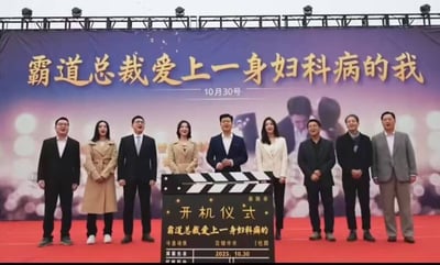 这张图片记录了一部网络剧集的开机仪式现场，舞台铺设红色地毯，后方背景板为紫粉渐变配色，上面以金色大字标注了该剧的剧名《霸道总裁爱上一身妇科病的我》，还标注了日期10月30号，背景板下方还有模糊的言情向人物剪影；共有9名穿着正装的主创人员整齐站在红毯上，大多面带笑容，台前放置了一块电影场记板样式的立牌，上面写有“开机仪式”字样，再次标注了该剧剧名，还印有2025.10.30的日期信息。该剧剧名设定十分猎奇，和常规甜宠言情剧的剧名风格反差极大，充满荒诞感。