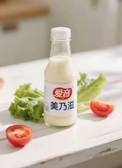这是一张爱音品牌美乃滋的产品宣传图，透明玻璃瓶内装着奶白色的美乃滋酱料，瓶身标签上清晰印有红色的“爱音”品牌标识与黑色的“美乃滋”品名。画面中还搭配了新鲜的生菜叶和切开的小番茄，摆放在浅色的木质桌面上，整体风格清新自然，直观展现出这款酱料适合搭配轻食、沙拉的使用场景，没有双关、谐音梗等内容。