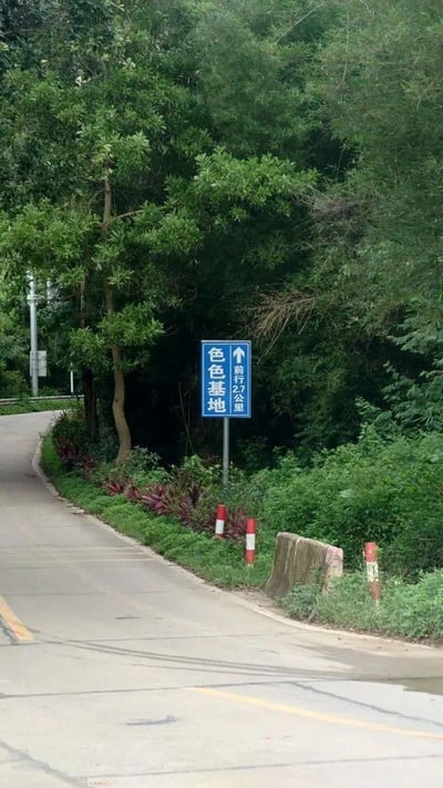 这是一条乡间的水泥道路，路边植被茂密，立着一块蓝色的道路指示牌，指示牌上写着“色色基地”，箭头指向前方，标注距离还有27公里。道路旁有红白相间的警示桩和路障，整体环境绿意盎然。
注：“色色基地”的名称带有谐音趣味，容易引发调侃联想。