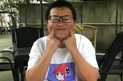 图中是一位戴着黑框眼镜的男生，身处户外休闲桌椅区域，他双手抬起，用手指将脸颊向上推挤，做出搞怪又带点可爱的表情。男生穿着印有红发动漫角色的白色T恤，整体画面氛围轻松诙谐，充满生活化的趣味感。