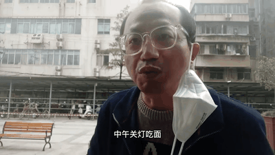 一位戴着眼镜的中老年男子站在居民楼前的广场上，口罩挂在脖子上，表情平静地重复说着'中午关灯吃面'。背景中可以看到多层居民楼、长椅和停放的自行车，整体环境呈现出普通城市生活区的景象。男子的话语直接引用了股市经典梗'关灯吃面'，通过真人实景的方式演绎了这一表达失意情绪的流行语。