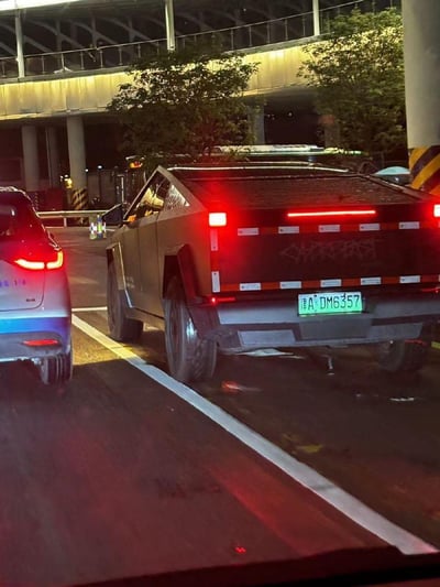 夜间道路场景下，一辆灰色特斯拉Cybertruck正行驶或停靠，车尾红色尾灯亮起，车牌号为“津A·DM6357”。车辆后方有明显涂鸦痕迹，左侧并排行驶着一辆银色轿车。背景可见立交桥结构、路灯及绿化树木，整体呈现出城市夜间交通的真实氛围。