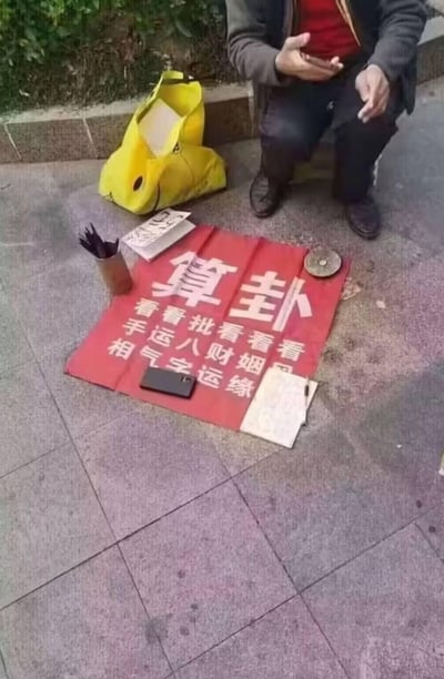 这是一张户外人行道的实拍图，地面铺着红底白字的「算卦」服务布，标注了看手相、看运气、批八字、看财运、看姻缘等服务项目，旁边摆放着签筒、相关书本、罗盘等算卦工具，一位男子蹲在布的旁边，做出手势，看起来是提供算卦服务的从业者。这类街头算卦属于封建迷信范畴，没有科学依据。