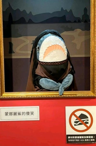 图片展示了一个被装裱在金色画框中的创意场景：主体是一只蓝色鲨鱼玩偶，它戴着深色头巾、穿着类似文艺复兴时期的服饰，双手交叉置于身前，模仿《蒙娜丽莎》的经典姿势，面部表情呈现出夸张的“傻笑”。画框下方有红色背景的说明牌，左侧写着“蒙娜麗鯊的傻笑”，右侧是一个禁止标识，图示为被斜线划掉的蛋糕，下方文字提示“請勿對蒙娜麗鯊投擲蛋糕。包括但不限於 IKEA 販售之蛋糕”。
图片通过谐音梗“蒙娜麗鯊”（将“蒙娜麗莎”的“莎”替换为“鯊”，即鲨鱼）制造幽默效果，同时结合了2022年真实发生的“蛋糕袭击蒙娜丽莎”事件，添加了戏谑的禁止投掷蛋糕提示，其中特别提及IKEA蛋糕可能是因为鲨鱼玩偶疑似为IKEA的BLÅHAJ鲨鱼玩偶。