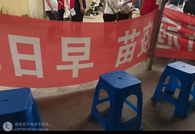 这是一张户外实拍的静态图，画面核心是一条被倒置悬挂的红色疫苗宣传横幅，原本的标语“新冠疫苗 早日接种”因悬挂方向错误，文字完全颠倒，呈现出“日早 苗疫冠新”的怪异组合，制造出滑稽的乌龙效果。画面下方摆放着三个蓝色塑料方凳，背景中能看到几位身着红白服饰的人员，场景推测为一处户外疫苗接种点的等候区域，整体氛围轻松搞笑，是现实中出现的趣味失误场景。