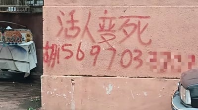 这是一张街头实拍的静态照片，画面主体是浅粉色的水泥外墙上用红色喷漆喷涂了手写风格的内容，上半部分是四个大字“活人梦死”，下半部分是姓氏“胡”加上手机号码1569703，号码末尾数位做了马赛克模糊处理。墙面左侧可以看到搭着遮阳布的贵州糯米饭售卖小摊的局部，右下角露出了机动车的车头部分，整体是违规喷涂在公共墙面上的小广告，因文字内容诡异反常具有很强的猎奇感。