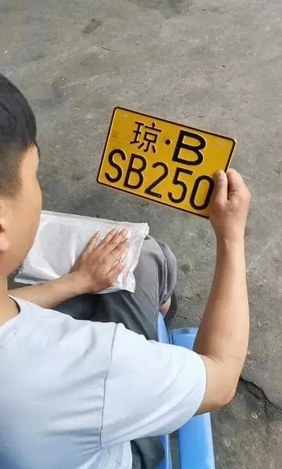 一名身穿浅蓝色短袖的男子坐在蓝色板凳上，手中举着一块黄色车牌，车牌上印有黑色文字“琼·B SB250”。背景为灰色水泥地面，男子左手放在腿上的白色塑料袋上。车牌号码“SB250”因“SB”在中文网络语境中常被用作调侃他人愚笨的俚语，“250”也有类似含义，二者组合形成极具幽默效果的车牌号码。