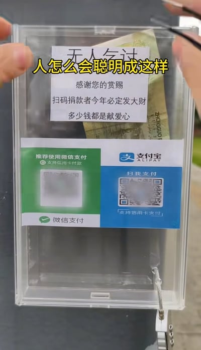 这是一张实拍的竖版图片，画面中有人手持一个带锁的透明亚克力募捐盒，盒子内已经放置了一张纸币。盒子上半部分贴有打印的“无人乞讨”说明文字，内容为“感谢您的赏赐，扫码捐款者今年必定发大财，多少钱都是献爱心”；下方并排张贴了微信支付和支付宝的官方样式收款码，均标注支持信用卡付款。图片上方叠加了黄色描黑边的醒目吐槽字幕“人怎么会聪明成这样”，整体调侃了将传统乞讨行为数字化、无人化的新奇操作，充满幽默感。