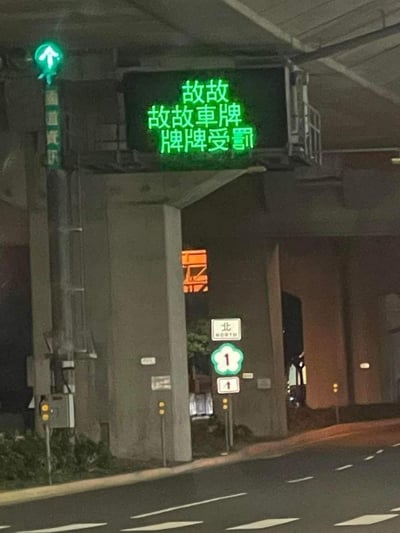这是一张夜晚拍摄的台湾国道场景图，画面上方的交通电子显示屏因故障，将原本的「故障車牌 牌牌受罰」提示语显示为「故故 故故車牌 牌牌受罰」，重复的「故故」形成了魔性的叠词效果，意外制造出搞笑感。画面中还可见向上通行的绿色指示灯、国道1号北向的道路标志，整体是真实的道路设施故障现场，充满趣味。
**趣味点说明**：原本的提示语是告知故障车牌会受罚，故障后文字重复，让「故」字叠用，形成类似卖萌的魔性表述，自带笑点。