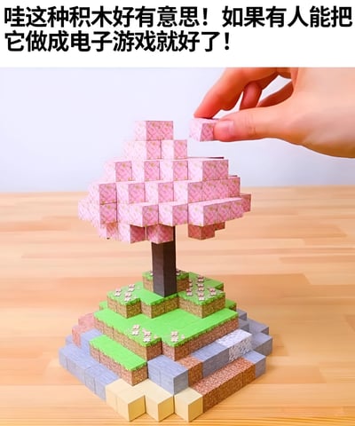 图片展示了一只手正在将粉色积木块添加到一个类似樱花树的积木结构上。树的底部是绿色平台，整体造型可爱。图片上方配有文字：'哇这种积木好有意思！如果有人能把它做成电子游戏就好了！'，表达了希望这种积木玩法能被开发成电子游戏的愿望。积木的方块风格让人联想到《我的世界》(Minecraft)等沙盒建造游戏。