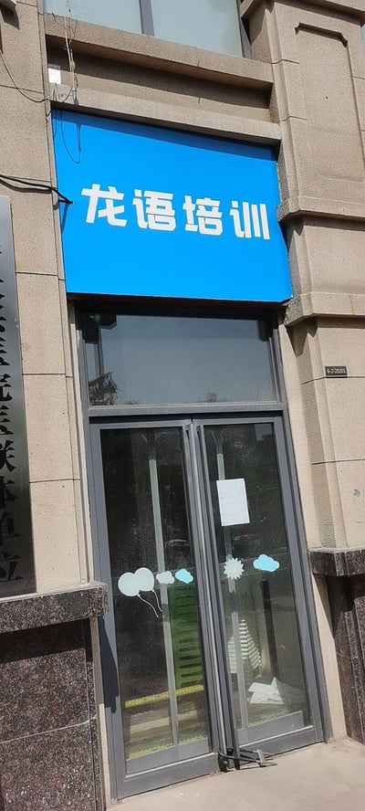 这是一张竖拍的线下培训机构门店照片，建筑外墙为浅棕色石材，上方悬挂一块蓝色招牌，印有白色的“龙语培训”字样。下方是灰色边框的玻璃双开门，门上贴有气球、云朵样式的可爱装饰贴纸，门右侧墙面有门牌号“6-2-20201”，左侧有一块部分可见的深色竖牌，上面露出白色的“医联体单位”字样。整体呈现出日常的临街商铺门头场景，无双关、谐音梗或暗示类内容。
