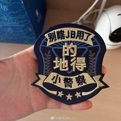 这是一只手拿着一枚警徽样式的刺绣布贴，布贴以藏蓝色为底色，搭配米黄、棕、蓝的刺绣线，设计成类似警察徽章的复古样式。布贴上的刺绣文字为“别瞎JB用了”“的地得”“小警察”，用幽默戏谑的方式吐槽中文里「的、地、得」这三个结构助词被乱用的普遍现象，以“小警察”的设定调侃要规范使用这三个易混淆的助词。
