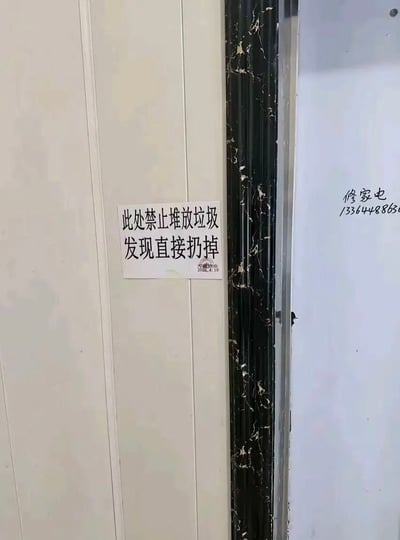 这是一张实拍的居民楼道墙面照片，米白色的竖纹墙面上贴着一张白底黑字的A4纸告示，告示内容为“此处禁止堆放垃圾 发现直接扔掉”，告示右下角盖有天鑫物业的印章，标注发布日期为2020年4月19日。告示右侧是带有白色纹理的黑色门框，相邻的另一处白色墙面上手写有修家电的联系电话小广告，号码未显示完整。