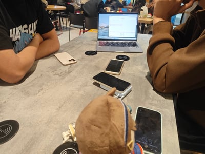 咖啡馆内的访谈视频拍摄现场：两人围坐在餐桌旁，桌上摆放着打开的笔记本电脑（屏幕显示疑似访谈内容界面）、多部智能手机、无线充电器及一个毛绒玩具。左侧人员穿着印有“HATSUNE”字样的黑色T恤，双臂交叉；右侧人员身着棕色外套，手托脸颊。背景可见其他顾客和桌椅，整体氛围 casual 且专注，展现了户外访谈拍摄的真实场景。