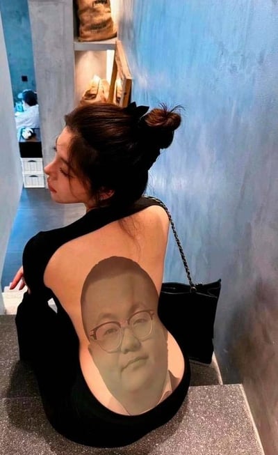 一位女子背对镜头站立，穿着一件设计独特的黑色露背装。令人惊讶的是，在她的后背上印着一张清晰的男性面部图像，该男性戴着眼镜，表情平静正视前方。女子将头发梳成一个发髻，戴着黑色蝴蝶结，肩上挎着一个黑色链条包。背景是一个室内空间，有蓝色的墙壁和一些摆放物品的架子，远处还有另一个人坐在那里。这种创意穿搭设计制造出一种视觉反差和幽默感，吸引观者注意力。