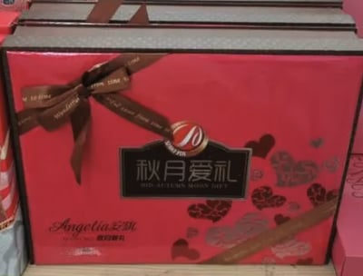 这是一款安琪（Angelina）品牌的中秋月饼礼盒，包装以鲜艳的红色为主色调，搭配印有英文祝福语的棕色装饰丝带，丝带系成蝴蝶结样式增添精致感。包装正面设有黑色标牌，清晰标注“秋月爱礼”及英文“MID-AUTUMN MOON GIFT”，上方带有安琪月饼的品牌标识，左下角印有“Angelina安琪”字样，同时包装上点缀着多组深浅不一的爱心图案，整体风格温馨浪漫，契合中秋送礼的温馨氛围。