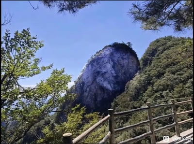 这是一张展示山间自然景观的摄影作品。画面中央是一块巨大的岩石山峰，表面呈现出深浅不一的灰色纹理，顶部覆盖着少量植被。山峰周围环绕着茂密的绿色树木，前景中可以看到一段木制围栏，可能是观景台的设施。天空呈现出清澈的蓝色，没有明显云层，整体画面给人一种宁静、自然的感觉。