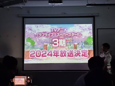 这是一张线下活动现场的翻拍照片，投影屏幕上展示了偶像动画《LoveLive!Superstar!!》TV动画第三季的官方消息，明确标注该季将于2024年播出，背景是动画中结丘女子高等学校的校门场景，现场有一位工作人员站立在屏幕旁，台下观众正用电子设备记录这一资讯，整体氛围充满粉丝对新动画的期待感。