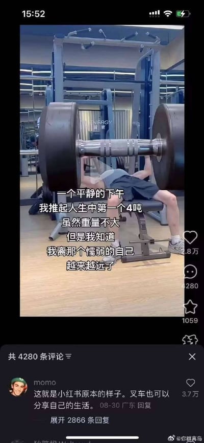 图片为小红书视频截图，画面中一名健身者在健身房卧推一个体积异常巨大的杠铃，杠铃片尺寸远超现实常理。图中叠加文字写道：“一个平静的下午 我推起人生中第一个4吨 虽然重量不大 但是我知道 我离那个懦弱的自己 越来越远了”，通过夸张的“4吨重量”制造幽默效果，讽刺网络上常见的过度夸大个人经历的现象。下方评论区中高赞评论“momo”调侃道：“这就是小红书原本的样子。叉车也可以分享自己的生活。”进一步强化了对平台夸张文化的戏谑。