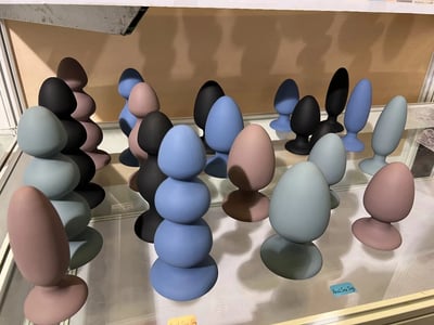 展示柜中陈列着多组色彩丰富的蛋形摆件，涵盖黑色、蓝色、粉色、灰色等多种哑光色调。摆件形态简约，部分为单个独立放置，部分呈堆叠状组合，整齐排列在透明展示柜内的白色台面上，背景为米色墙面，整体风格现代简约，适合作为家居装饰或设计展览展示品。