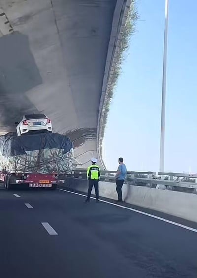 在一条高架桥梁路段，一辆盖着篷布的大型货运挂车，因上层装载的白色本田轿车高度超出限高，卡在了桥梁底部无法通行。一名身着反光警服的交警和一名疑似货车司机的男子站在护栏边，正在现场处理这一突发的交通状况。