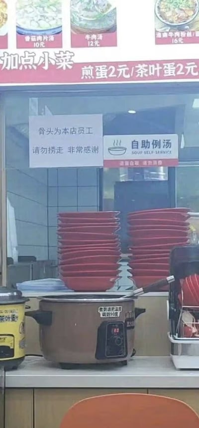 图片展示了一家餐馆的取餐窗口场景。窗口上方张贴着菜品图片及价格，包括香菇肉片汤10元、牛肉汤12元、逍遥牛肉粉丝汤16元等，下方红色横幅标注“加点小菜 煎蛋2元/茶叶蛋2元”。窗口内侧玻璃上贴有两张告示，左侧为“骨头为本店员工 请勿捞走 非常感谢”，右侧为“自助例汤”及英文“SOUP SELF-SERVICE”。因“骨头为本店员工”表述存在歧义，初看易让人误以为是“员工的骨头”，引发搞笑误会，实际应为店家提示汤中用于熬汤的骨头为店内自用，请勿顾客捞走。