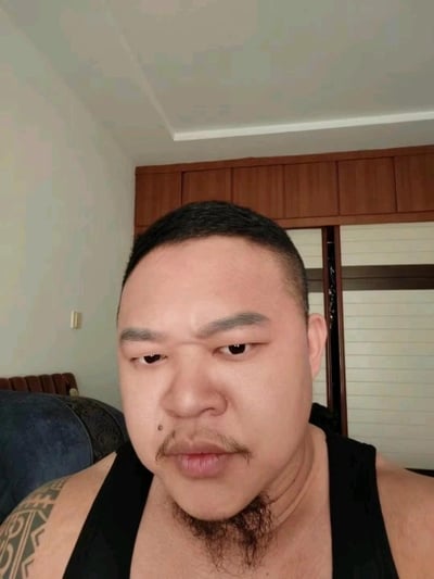 这是一张室内环境下的真人自拍图，图中男子穿着黑色背心，留着上唇小胡+下巴络腮胡的特色胡须造型，左手臂带有图腾样式纹身。他眉头微皱，左眼正常睁开，右眼半睁，呈现出搞怪又带点疑惑的表情，正对着镜头拍摄。背景可见居家空间，包含木质衣柜、沙发和暖气片，整体氛围生活化。图中没有双关、谐音梗或暗示内容。