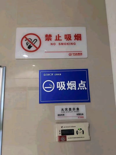 这张图片拍摄于辽阳文圣万达广场的浅米色瓷砖墙面，从上到下依次排布内容：最上方是一块白色底的禁止吸烟标识牌，左侧是红圈斜杠加香烟的禁止吸烟标准图案，右侧印有红色中文“禁止吸烟”与对应英文“NO SMOKING”，右下角印有万达集团的品牌标识；其下方紧邻一块蓝色底的标识牌，左侧是带香烟烟雾的吸烟图标，右侧印有白色大字“吸烟点”，左上角标注“万达广场 辽阳文圣”；再往下是白色底的“火灾显示盘”提示标识，标注了“消防专用 非警勿动”；最下方是实际的北大青鸟品牌火灾显示盘消防设备。同一个墙面的紧邻位置同时设置“禁止吸烟”和“吸烟点”两块相反功能的标识，形成了矛盾滑稽的观感。