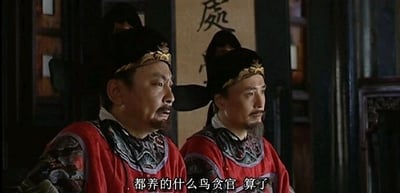 这是电视剧《大明王朝1566嘉靖与海瑞》的剧照截图，画面呈现了室内场景中的两位明代官员，左侧为郑泌昌，右侧为何茂才，二人皆身着带有精致补子的红色官服，头戴明代官帽。左侧的郑泌昌面露愤懑，眉头紧皱，嘴巴微张，正在抱怨“都养的什么鸟贪官 算了”，情绪激动；右侧的何茂才则面色凝重，眼神看向画面右侧，神情严肃，整体场景传递出官场人物面对乱象时的无奈与愤懑情绪，背景的书法牌匾也增添了古雅的官场氛围。