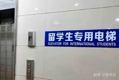 这是一张室内电梯等候区的实拍照片，画面右侧的浅色瓷砖墙面上，悬挂着一块蓝底白字的标识牌，上方是简体中文“留学生专用电梯”，下方对应英文翻译“ELEVATOR FOR INTERNATIONAL STUDENTS”。画面左侧可以看到部分打开的电梯门和金属质感的电梯呼叫按钮面板，整体场景简洁，是常见的楼宇电梯区域设置。该标识的特殊性在于为留学生设置了专用电梯，这种差异化的设施配置容易引发公众对于资源分配相关的讨论。