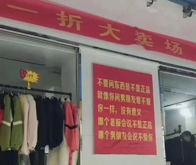 这是一张线下实体服装店的实拍图，店铺上方悬挂着红色的“一折大卖场”横幅，店内挂着多件标价199元的针织类外套，颜色多样，有绿色、米色、黑色、粉色等。店铺墙面贴着一张醒目的红色宣传标语，用生活化的类比来回应顾客对商品是否为正品的疑问：将“问老板商品是不是正品”和“问男朋友爱不爱你”放在一起类比，指出两者的答案都是显而易见的，老板不会说自己的商品不是正品，就像男朋友不会说不爱你，用幽默扎心的接地气风格来做店铺促销宣传，右下角还能看到一位似乎在看衣服的顾客。