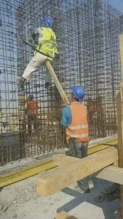两名建筑工人在施工现场进行作业。一名身穿黄色安全背心的工人正站在木板上攀爬密集的钢筋架，另一名身穿橙色安全背心的工人则在地面扶着这块充当临时梯子的木板。两人均佩戴蓝色安全帽，背景中可以看到更多的钢筋结构和隐约的其他工人身影，展现了建筑施工中的真实工作场景。