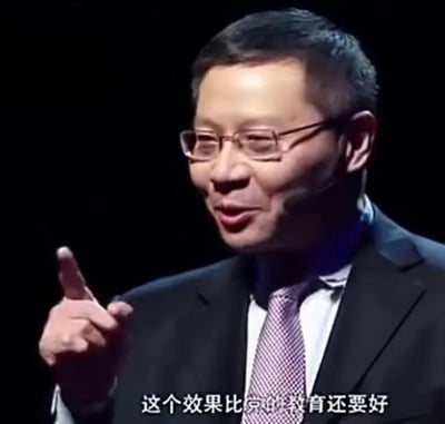 这是一张真实人物张维为的单帧梗图，画面中张维为佩戴细框眼镜，身穿黑色西装搭配淡紫色格纹领带，面带笑意神态轻松，抬起右手食指指向前方正在发言，画面底部配有白色字幕“这个效果比北京的教育还要好”
