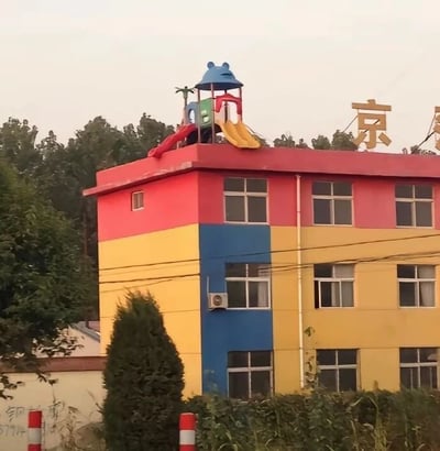 这是一张展示幼儿园建筑的照片。建筑主体为多层结构，外墙采用鲜艳的色彩搭配，顶部楼层为红色，中间楼层为黄色，并有一条蓝色的垂直装饰条。建筑屋顶上安装了一套儿童游乐设施，包括带有蓝色顶棚的卡通造型结构，以及红色和黄色的滑梯。背景中可以看到茂密的树木和一些电线杆，建筑前方有绿色的灌木丛和红白相间的路障。屋顶右侧有一个金色的汉字“京”，可能是该幼儿园名称的一部分。整体画面给人一种童趣、活泼的感觉，适合儿童活动场所的氛围。
