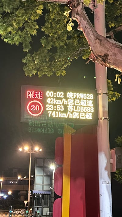 这是一张夜晚户外拍摄的照片，画面核心是道路旁的电子测速显示屏：左侧红色标识明确限速20km/h，屏幕上列出两辆车的超速记录，其中皖FRW628以42km/h超速属于正常范畴，但苏LDB688被显示车速达741km/h，显然是测速设备出现故障，这个违背现实的离谱车速形成了搞笑的反差效果。背景可见行道树、照明路灯和城市建筑物，整体是夜晚的城市道路场景。