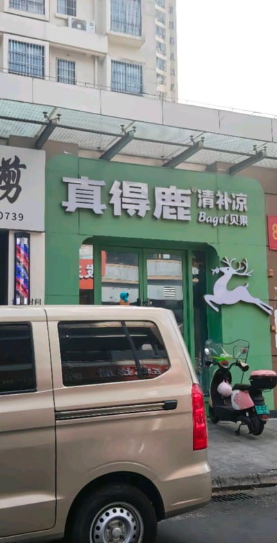 图片展示了一家名为“真得鹿清补凉贝果”的店铺门面。店铺外墙为绿色，正上方有白色大字招牌，左侧是中文“真得鹿 清补凉”，右侧标注英文“Bagel贝果”，门旁有一个白色的跳跃鹿图案logo。店门口停放着一辆米色面包车和一辆粉色电动车，背景可见高层居民楼，整体呈现出日常街头的店铺景象。