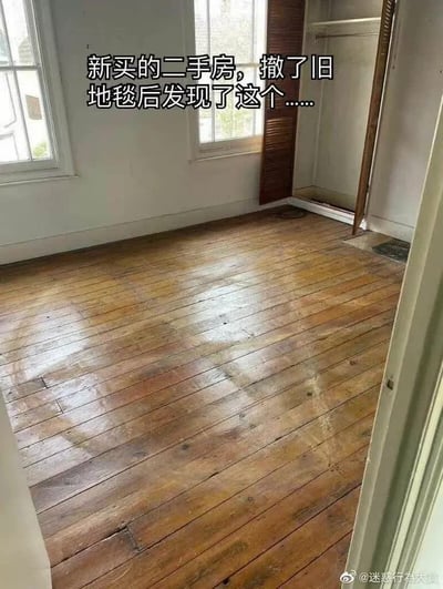 这是一张带文字注解的实拍单图，展示了新买的二手房的空房间，房间墙面为白色，设有两扇窗户采光，右侧带有开放式壁柜空间，从门口视角拍摄。图片上方配有悬念式文字“新买的二手房，撤了旧地毯后发现了这个......”，撤掉旧地毯后露出了房间原本铺设的长条实木地板，大部分地板保存完整，仅在靠近壁柜入口处有一小块区域的地面材质和原木地板不同，整张图通过这种留白悬念的方式引发观者好奇，容易让人联想到恐怖或意外的发现，实际图中仅露出了原本的旧木地板，右下角带有微博账号@迷惑行為大賞的水印。