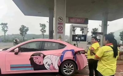 加油站场景中，一辆粉色轿车正在加油，车身侧面贴有大型动漫女孩贴纸。贴纸中的女孩有着黑色长发，表情惊讶，身穿白色上衣和蓝色背包，呈现出趴在车身上的姿态。车旁有两位穿着黄色工作服的加油站工作人员，正面向轿车站立，似乎在观察车身上的贴纸。背景可见加油站的加油机，上方显示“92# 95#”字样，周围有禁止吸烟等警示标识，远处有树木和模糊的山景。